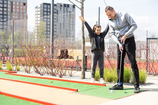 Sharing with golf experience. Cheerful young man teaching his daughter to play mini golf at the day time. Concept of friendly family.