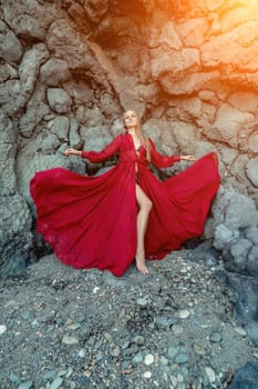 Red dress rocks woman. A blonde with flowing hair in a long flowing red dress stands near a rock of volcanic origin. Travel concept, photo session at sea.