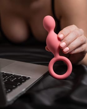 Woman holding pink anal beads next to laptop while lying on black sheet