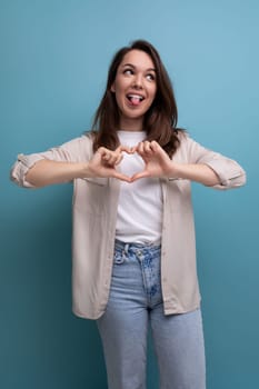30s woman with chic black hair below her shoulders in a beige shirt shows a heart with her hands.