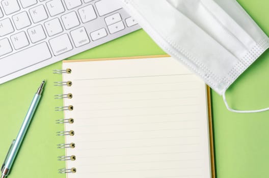 Open blank diary and keyboard computer with disposable face mask on green background.