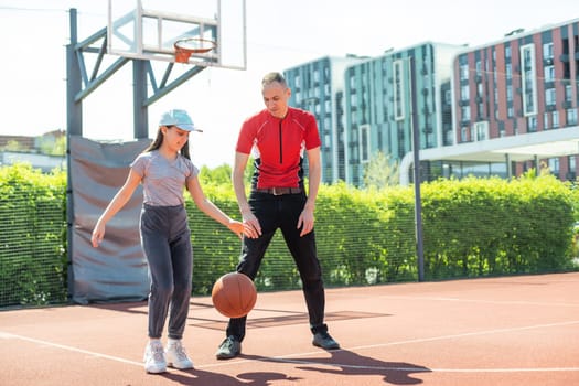 Happy father and teenage daughter playing basketball outside at court.