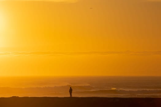 A lonely man stands by the sea at bright orange sunset. Mid shot