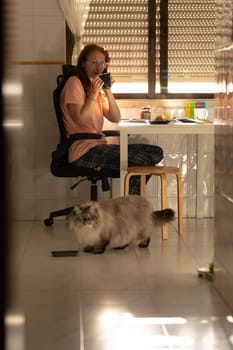 An adult woman sits at the kitchen table and drinks coffee - a cat sits next to her on the floor. Vertical shot