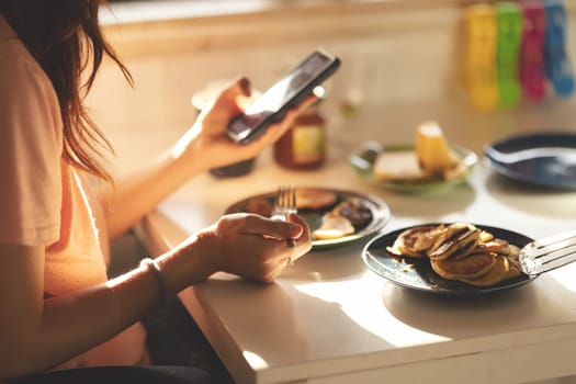 An adult business woman eats pancakes for breakfast and looks at her phone at the kitchen table. Mid shot