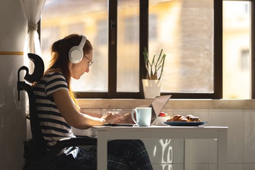 Adult woman freelancer in headphones working at the kitchen table. Mid shot