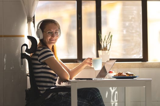 Smiling adult woman in headphones sitting by the kitchen table with a cup of coffee. Mid shot