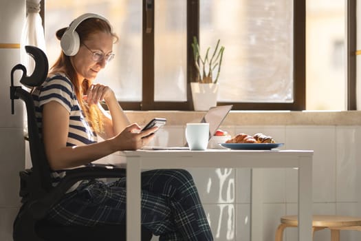 An adult business woman in headphones sitting by the kitchen table and looking at her phone. Mid shot