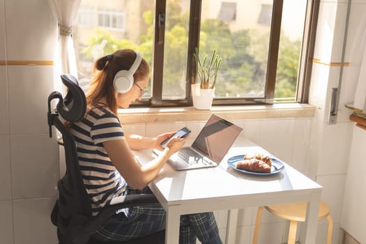 An adult woman freelancer in headphones sitting by the kitchen table and looking at her phone. Mid shot