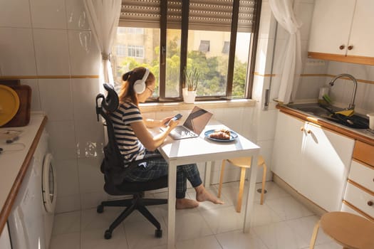 An adult business woman in headphones working at the kitchen table. Mid shot