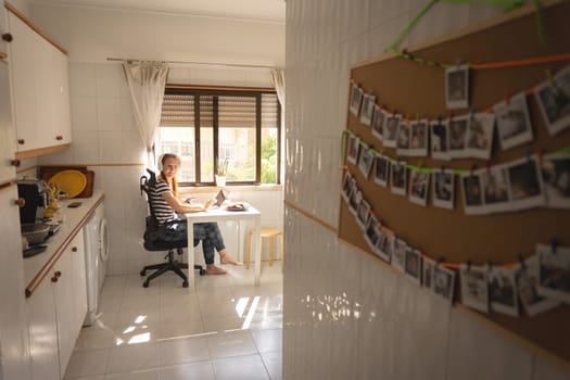 An adult business woman in headphones working at the kitchen and looking in the camera - a board with printed photos in the foreground. Mid shot