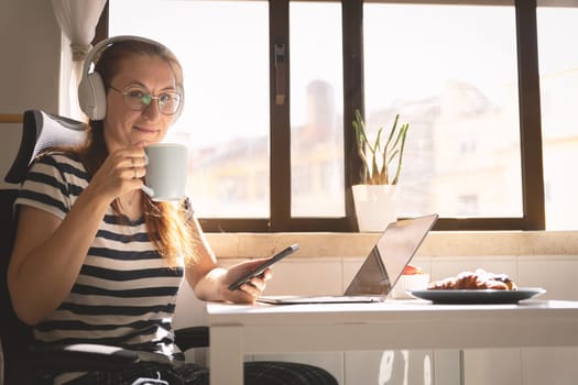 Smiling business woman in headphones sitting at the kitchen table and drinking coffee - looking in the camera. Mid shot
