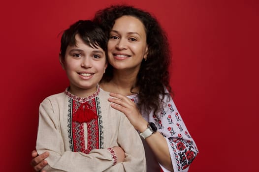 Beautiful woman, happy mother and her son wearing Ukrainian ethnic embroidered shirts Vyshyvanka, smiling looking at camera, isolated on red background. Ukraine Traditions Folklore Culture and Fashion