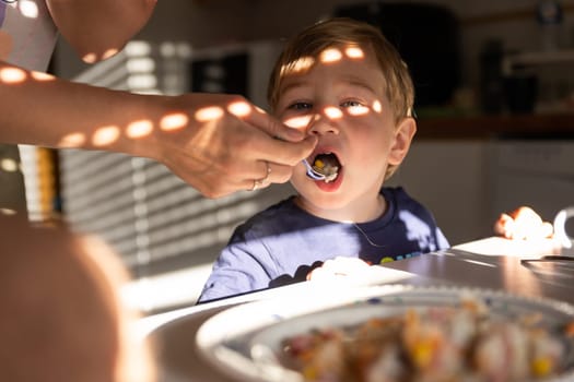 Mom feeds a boy with a spoon in the kitchen. Mid shot