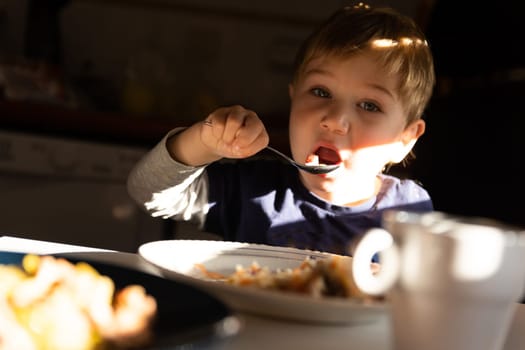 A little boy eats breakfast at the table - eats with a spoon and looks into the camera. Mid shot