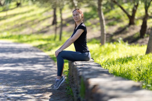 Adult smiling woman in sports clothes sits on the large stone curb and looking in the camera. Mid shot