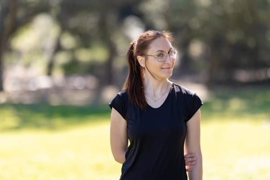 An adult smiling woman wearing glasses and sports clothes standing in the park. Mid shot