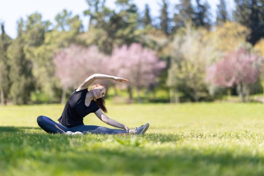 Yoga on nature - an adult woman sitting in lotus position and bending to the side with her leg stretched out. Mid shot