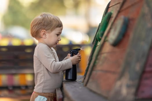 A little boy sprays water on a wooden boat from a spray gun on playground. Mid shot