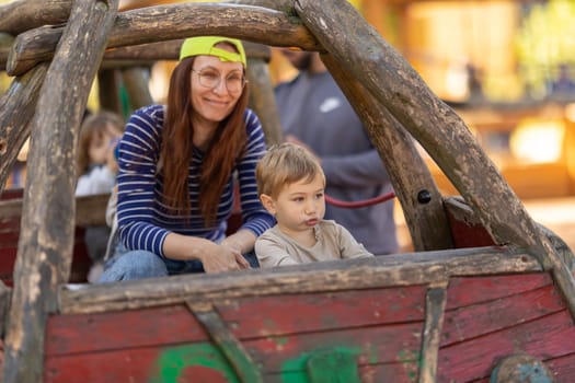A little funny boy and his smiling mother sitting in the wooden boat on the playground. Mid shot