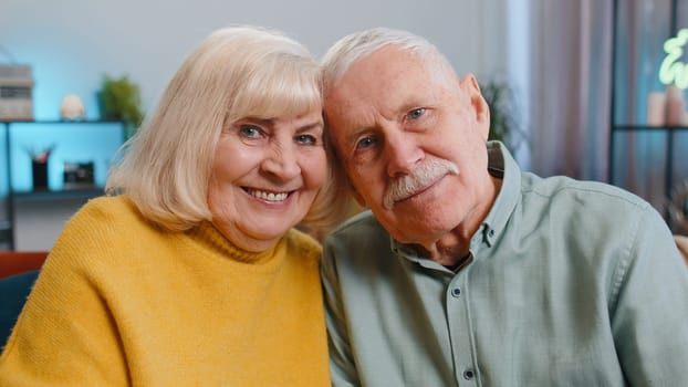 Happy old senior elderly family couple hugging, laughing, smiling looking at camera, healthy retired two grandparents husband wife. Grandmother grandfather embracing sitting on sofa at home, portrait