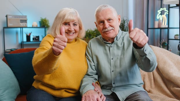 Like. Happy senior family grandparents man woman showing thumbs up, like sign positive something good, positive feedback at home. Elderly grandmother, grandfather couple sitting on sofa in living room