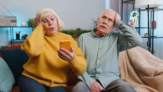 Senior family grandparents man woman use smartphone typing browsing, loses surprised by lottery results, bad news, fortune loss, fail at home. Elderly grandmother, grandfather couple on sofa in room