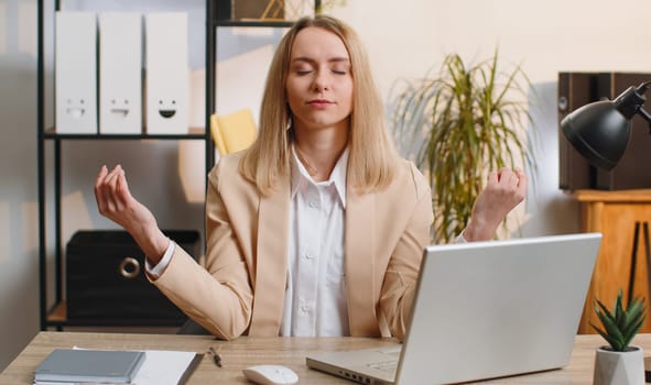 Young businesswoman working on laptop computer, meditating, doing yoga breathing exercise in lotus position at home office. Calm serene freelancer woman taking break. Busy occupation. Peace of mind