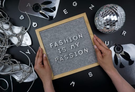 The quote fashion is my passion is laid out in the frame. There are shiny letters in the frame, shiny objects lie around - a disco ball, silver shoes, black earrings, letters made of rhinestones. A board with a glamorous inscription in female hands.
