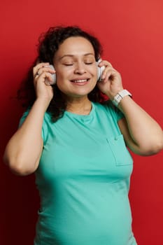 Close-up studio portrait of a happy pregnant woman with big belly, expressing positive emotions while listening to music on wireless headphones, isolated over red background. People Leisure Lifestyles