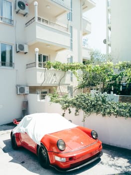Red Porsche 911 Carrera stands covered with a cloth near an apartment building. High quality photo