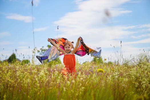 Portrait of little girl with Asian eyes and butterfly wings having fun and joy in meadow or field with grass, flowers on sunny summer day