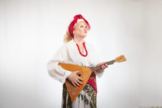 Cheerful funny adult mature woman solokha with musical balalaika. Female model in clothes of national ethnic Slavic style. Stylized Ukrainian, Belarusian or Russian woman poses in comic photo shoot