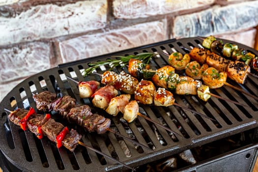 A lot of mini-kebabs of meat, fish, chicken, shrimp, vegetables on wooden skewers are fried on a small cast-iron grill.