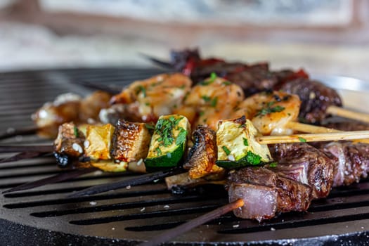 A lot of mini-kebabs of meat, fish, chicken, shrimp, vegetables on wooden skewers are fried on a small cast-iron grill.