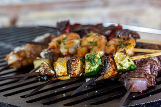 A lot of mini-kebabs of meat, fish, chicken, shrimp, vegetables on wooden skewers are fried on a small cast-iron grill.