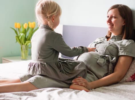 Attractive smiling pregnant woman with her little daughter are talking and spending time together at home. Lovely child touching mothers pregnant belly. Expecting mother sitting with her cute girl.
