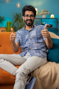 Like. Happy excited indian man looking approvingly at camera showing thumbs up, like sign positive something, good great news, positive feedback. Young hindu guy sitting on couch at home living room