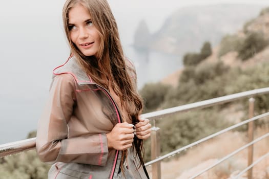 Woman rain park. Happy woman portrait wearing a raincoat with transparent umbrella outdoors on rainy day in park near sea. Girl on the nature on rainy overcast day