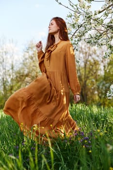 a happy, slender, sweet woman stands in a long orange dress in the tall grass near a flowering tree and happily smiling throws up the hem of her dress, looking away. High quality photo