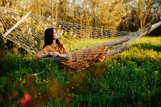 an elegant, slender woman is resting in nature lying in a mesh hammock in a long orange dress enjoying the rays of the setting sun on a warm summer day looking away. High quality photo