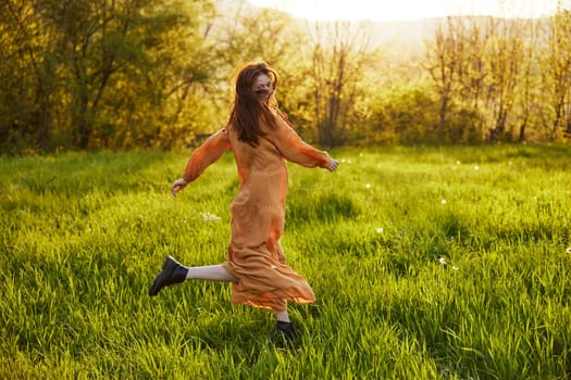 a happy woman in an orange dress illuminated by the rays of the setting sun is happily spinning in a green field, enjoying nature on a warm summer day. High quality photo