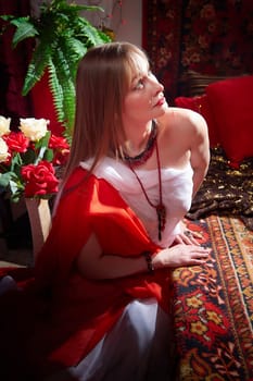 Beautiful European girl looking like Arab woman in red room with rich fabrics and carpets in sultan harem. Photo shoot of an oriental style odalisque. A model poses in sari as indian woman in india