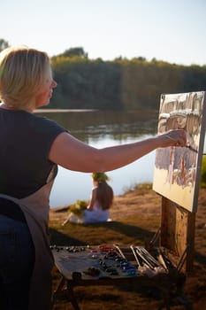 Adult female artist painting picture near water of river or lake in nature and girls in white sundress and flower wreath. Artist and models posing in holiday of Ivan Kupala in nature at sunset