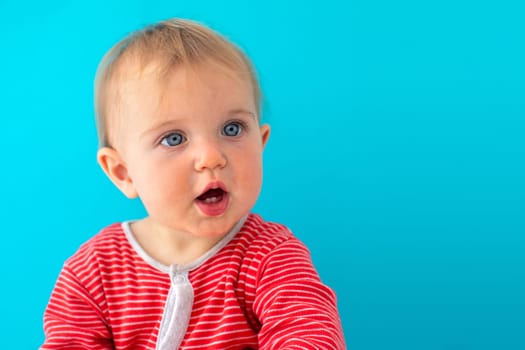 Sweet baby girl closeup portrait of child looks away surprised isolated on blue background, cute toddler with blue eyes in red