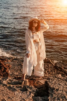 woman sea sunset. woman in a white pantsuit and hat is standing on the beach enjoying the sea. Happy summer holidays.