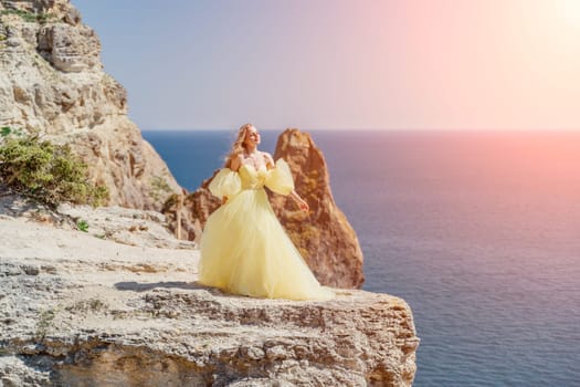 Woman yellow dress sea. Side view Young beautiful sensual woman in yellow long dress posing on a rock high above the sea at sunset. Girl in nature against the blue sky.