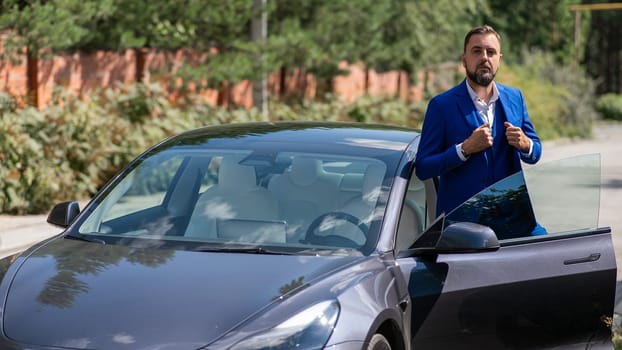 Caucasian bearded man in a blue suit gets out of a black electro car in the countryside in summer