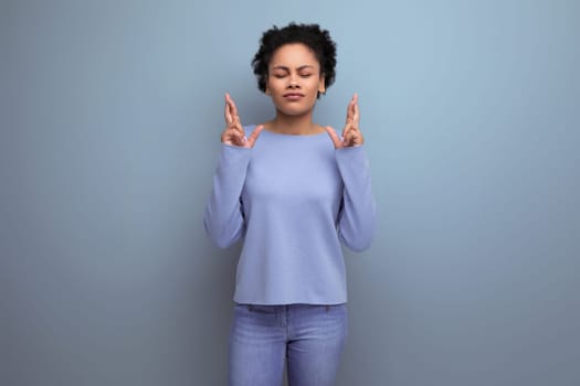 young pretty brunette woman with afro curls crossed fingers in hope on blue background with copy space.