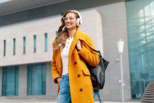Woman Wear Eyeglasses Listen Music Headphone Walking Outdoor City Street Dressed Stylish Yellow Coat And Black Backpack Smile Caucasian Female 30s Enjoy Podcast Or Audio Books Outside